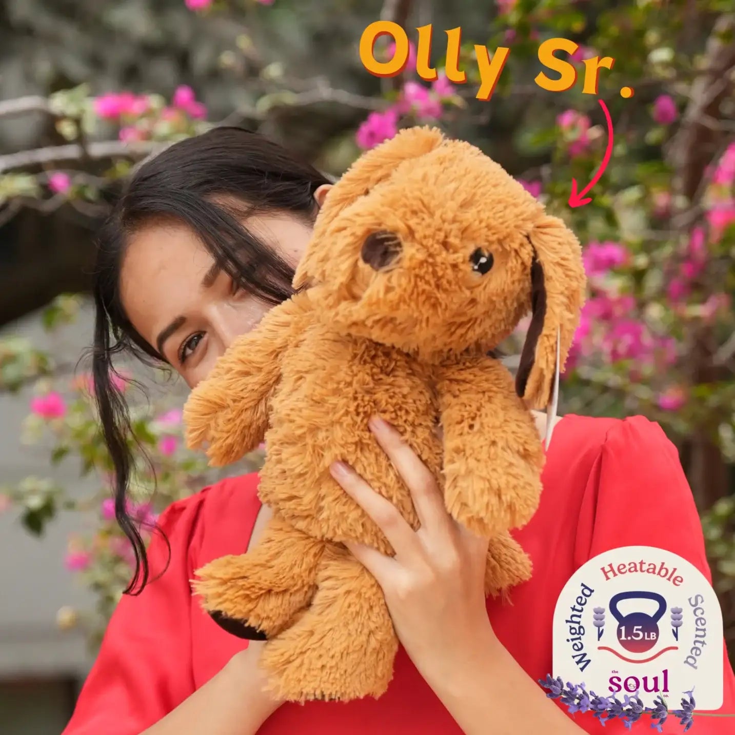 Woman holding a large brown weighted plush dog with text 'Olly Sr.', heatable and scented badge.